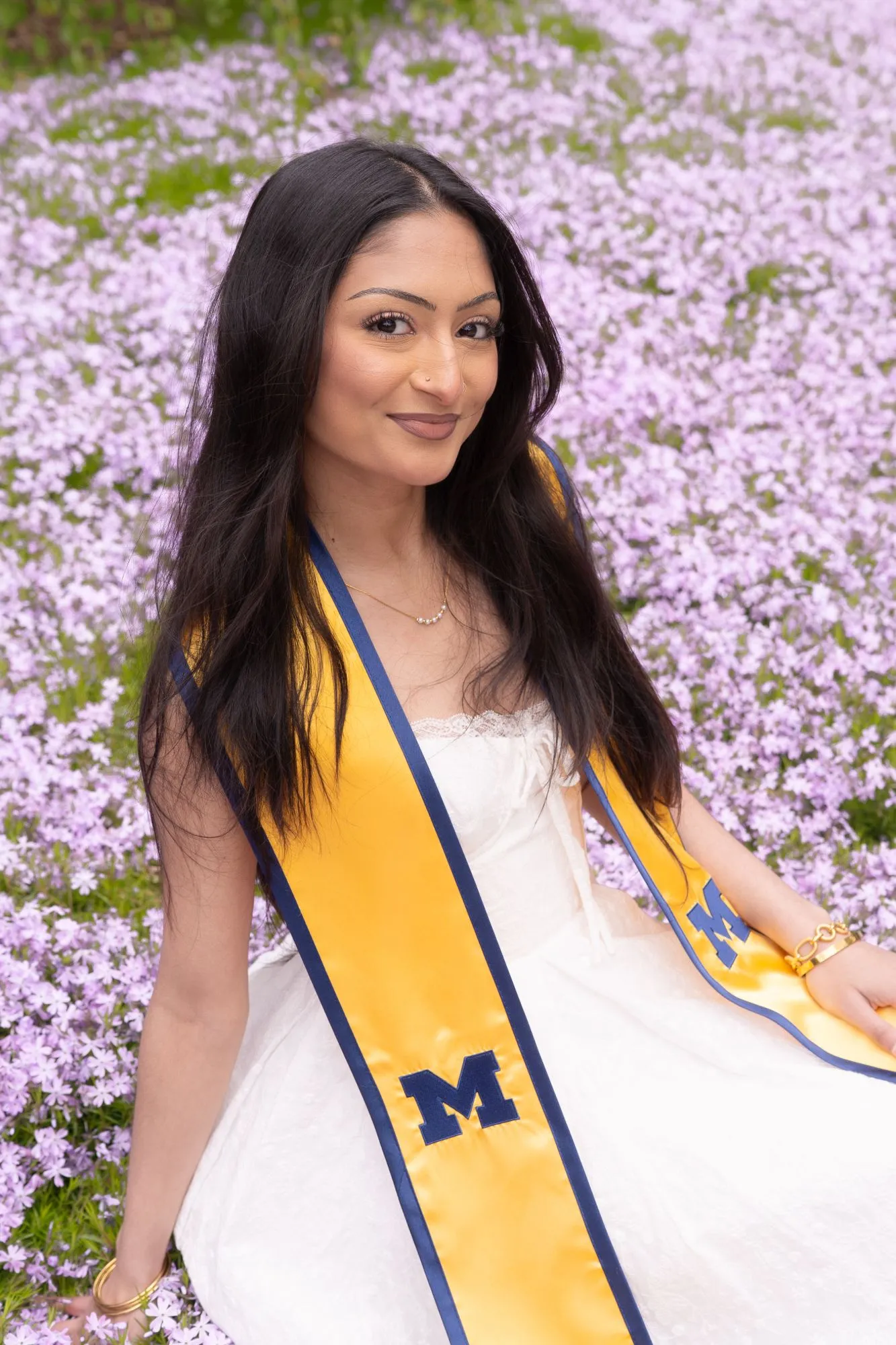 Tanya sitting in a field of lilac phlox flowers, smiling over her shoulder in a white dress and a maize-and-blue Michigan graduation stole.